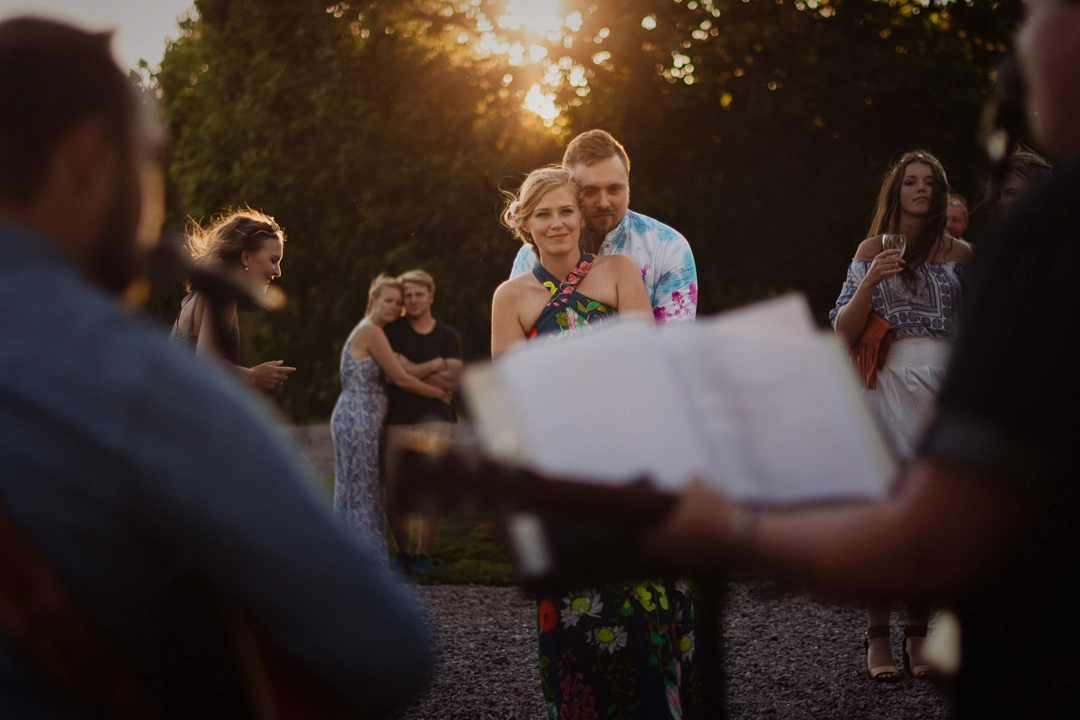 Timeless photo in the sunset of groom with arms wrapped around brides waist. They are dressed in 70's themed clothing enjoying the musicians performance. Wedding guests are enjoying the music in the background.
